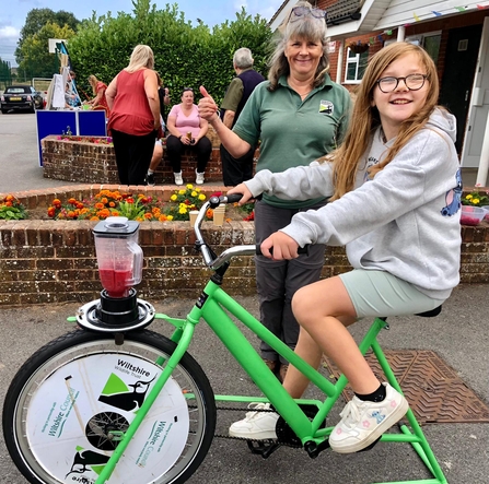 A young female on a static bike pedaling to create a smoothie, with an adult next to them and people in the background. 